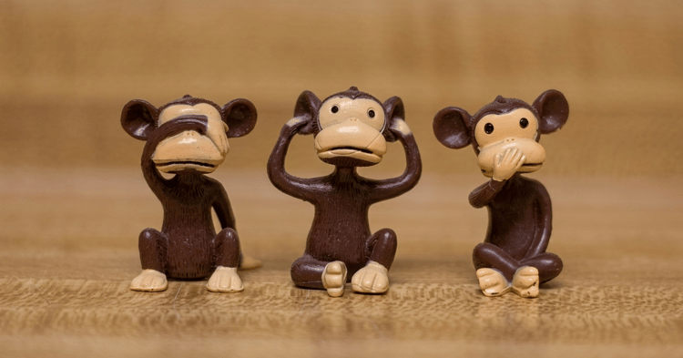 Three small dark brown monkey figurines sit on a wooden surface, each with hands covering different facial features