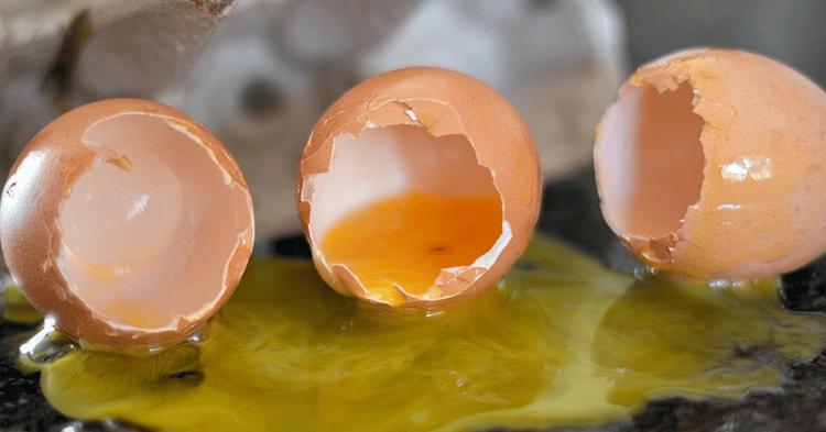 Three cracked eggshells sit on a dark surface, with the center shell containing a broken yolk and spilled yellow liquid