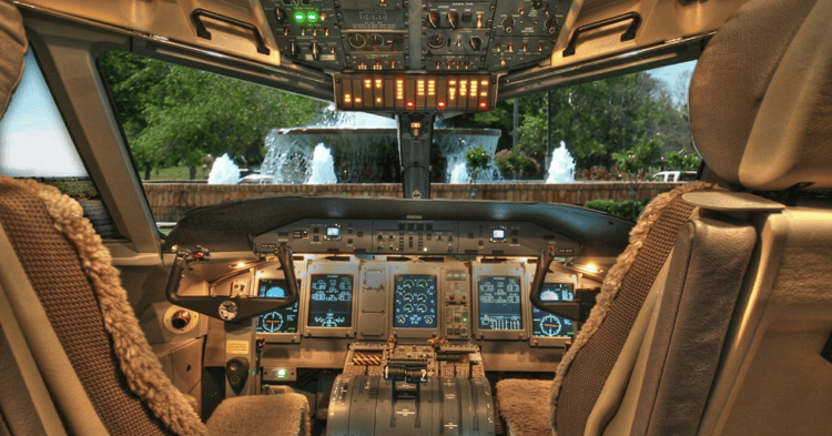 Airplane cockpit interior with instrument panels, a control yoke, and fuzzy-covered seats, overlooking trees and fountains through the windshield