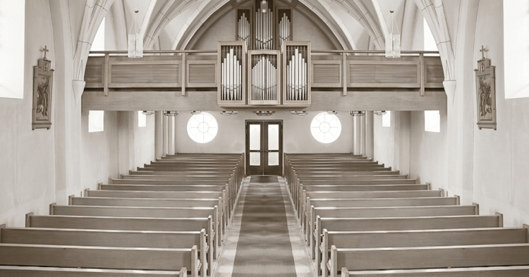 Black-and-white photograph of a church interior showing rows of wooden pews facing double doors, with a large pipe organ on an upper balcony