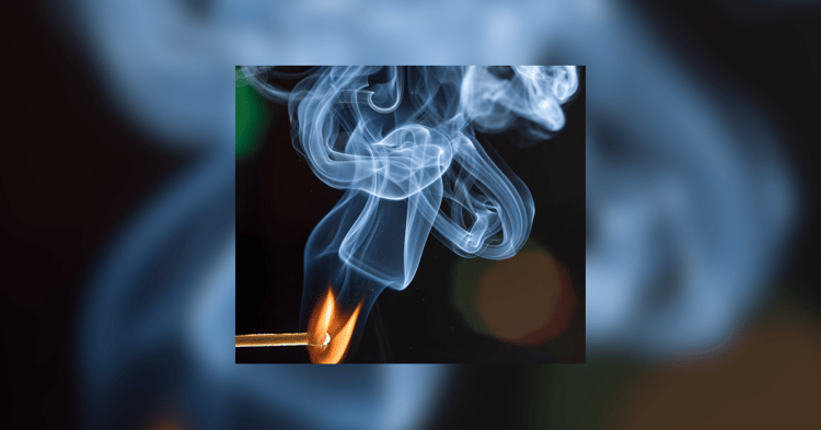 A lit matchstick with a bright flame and curling smoke rising against a dark, blurred background