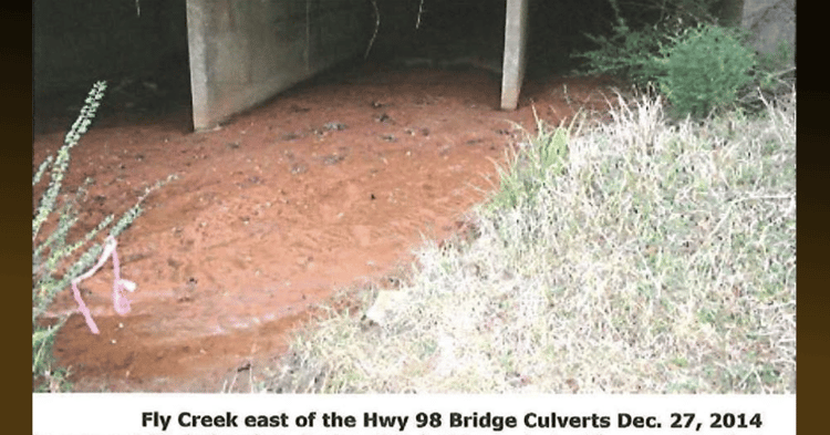 Muddy water flows under a concrete culvert in Fly Creek east of Hwy 98, with grassy banks. Caption: Dec. 27, 2014