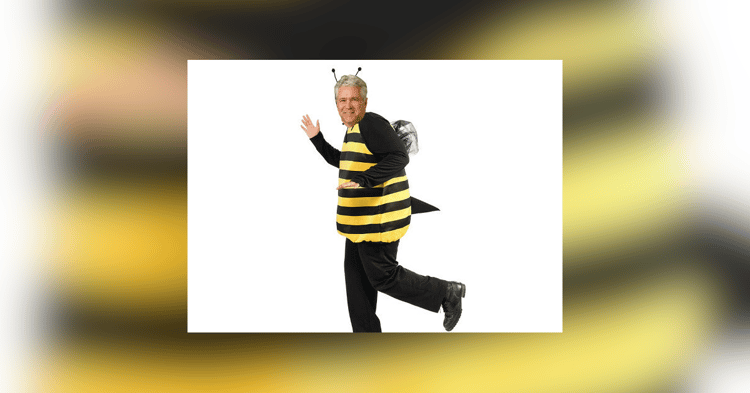 A man in a black and yellow striped bee costume with antennae and wing backpack waves while stepping forward against a white background