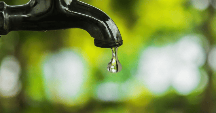 A close-up of a black faucet with a single water droplet hanging from its tip against a blurred green background