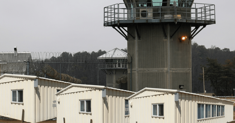 A correctional facility with a tall guard tower, barbed wire fencing, and white modular buildings under an overcast sky