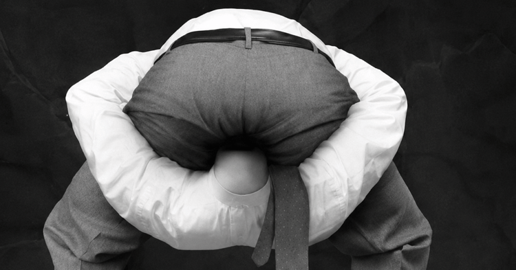 A black-and-white photo of a man in business attire bent forward with his head in his ass