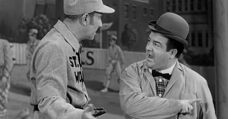 Two men in vintage attire converse outdoors; the man on the left wears a military uniform while the man on the right, in a bowler hat and bow tie, point...